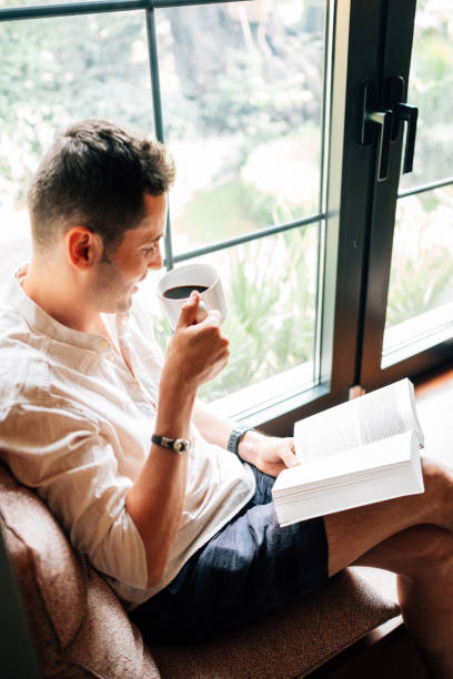 istock Young man reading a book in front of windowpane 1157614213