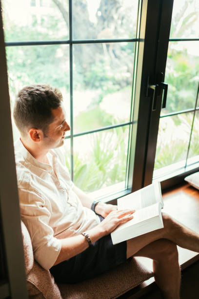 istock Young man reading a book in front of windowpane 1157614257