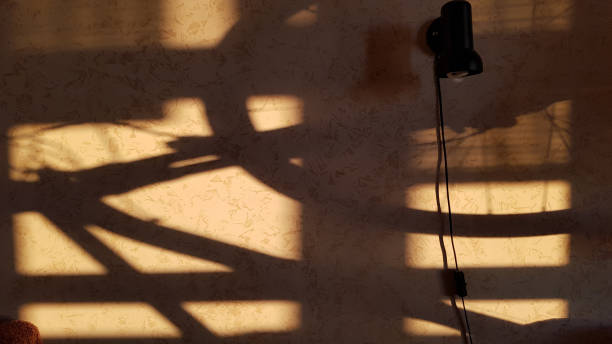 Shadows on stucco wall created by sunset light. Abstract background. Wall with black sconce lamp and dark crossed lines shadows with unclear contours. Light and shadow backdrop.