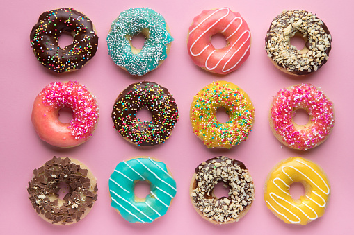 Stock photo showing elevated view of a blue plate containing an individual glazed ring doughnut decorated with white, pink, green, blue, yellow, and orange, hundred and thousand sugar sprinkles on blue background.