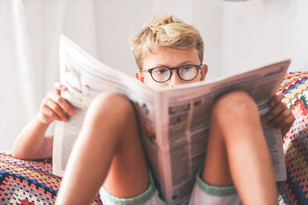 menino novo bonito que lê um jornal em casa. a criança loura lê a notícia em um jornal. geração z criança procurando notícias de última hora em uma revista de papel. informação interesse cultura conceito educacional - papery - fotografias e filmes do acervo