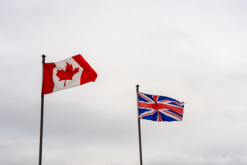 Flags of Canada and United Kingdom flapping in wind against cloudy sky.