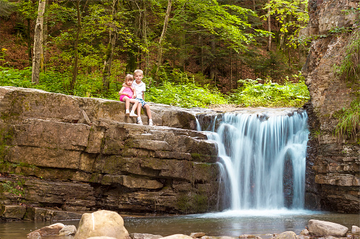 Two children boy and girl sitting near waterfall in forest