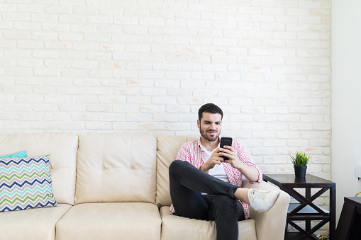 Male blogger sitting on sofa while checking his social media status on smartphone