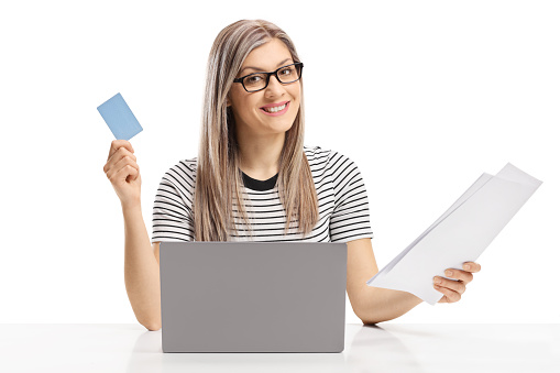 Young woman sitting with a laptop and holding a credit card and paper bills isolated on white background