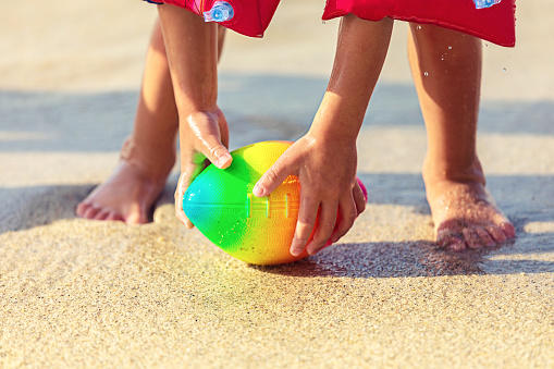 Baby feet walking on sand beach grabbing rugby ball - playful toddler wearing inflatable armbands hand holding ball from water in summer vacation - Child stand barefoot on sand grabbing ball