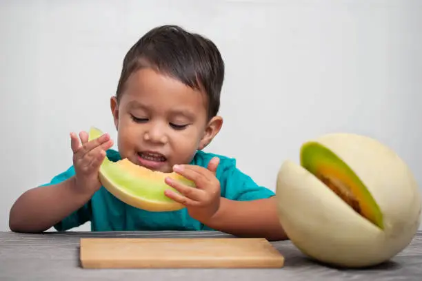 Photo of A little boy holding a slice of fresh melon fruit to his mouth that he has taken a bite out off, and enjoying the juicy sweet taste.