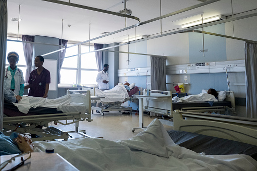 A large hospital ward with patients lying in their beds. Two doctors are doing their daily rounds checking the patients.