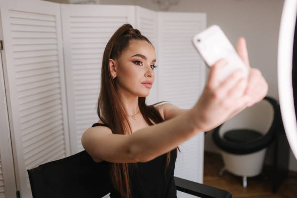 istock Young attractive model make a selfie after makeup in beauty studio. She use a ring lapm for photo. Background of white folding screen 1157712801