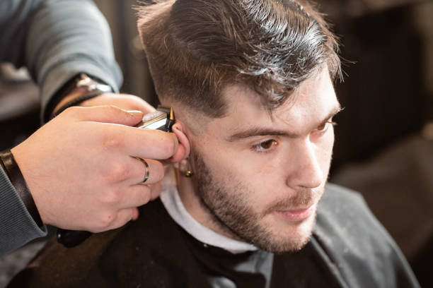 istock Barber work with clipper machine in barbershop. Professional trimmer tool cuts beard and hair on young guy in barber shop salon. 1157717689
