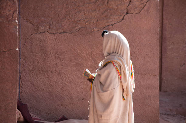 Ethiopian Pilgrim celebrating Orthodox Christmas Eve in Lalibela Lalibela, Ethiopia - 4 January, 2018: African Christian Woman in traditional white clothes standing with Bible in hand and praying near rock-hewn Bete Medhane Alem Church, side view ethiopian orthodox church stock pictures, royalty-free photos & images