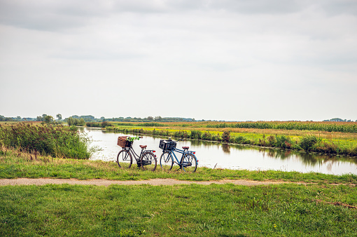 Women's and men's bicycle, each with a basket on the handlebars,  parked along the water of a stream in a Dutch polder. The owners are not visibly present in the area.