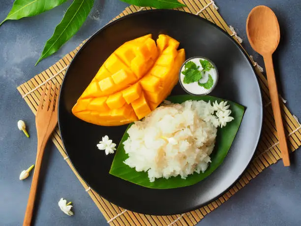 Mango sticky rice in a black plate, a famous Thai dessert for the summer season in top view.
