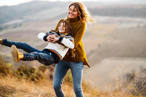 Young happy mother having fun while spinning her little daughter in autumn day on a hill. Copy space.
