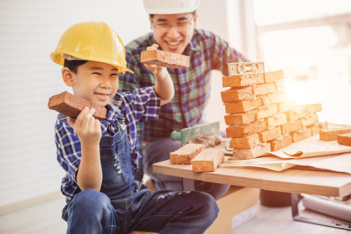 Father and little boy with bricks in hand and laughter. Man and little boy are enjoying playing as a construction engineer in the home. Love and relationships with the family.