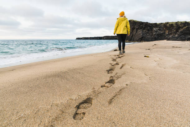 chica con impermeable amarillo caminando en la arena de la playa de skardsvik al atardecer, península de snaefellsnes, islandia - sand footprint track following fotografías e imágenes de stock