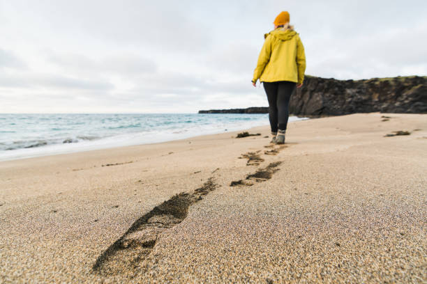 chica con impermeable amarillo caminando en la arena de la playa de skardsvik al atardecer, península de snaefellsnes, islandia - sand footprint track following fotografías e imágenes de stock