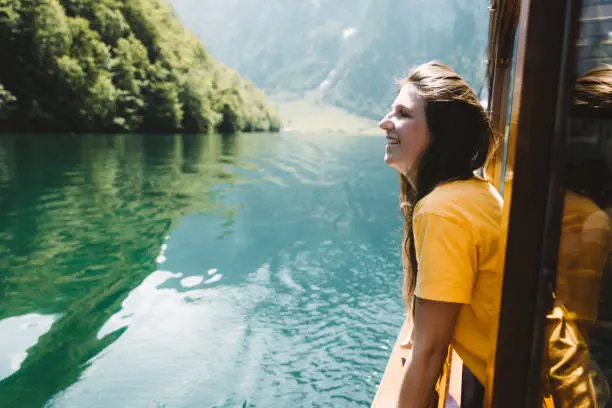 Young woman with long hair and yellow t-shirt riding by boat through beautiful turquoise mountain lake Konigsee with Alps view in Berchtesgaden, Bavaria, Germany
