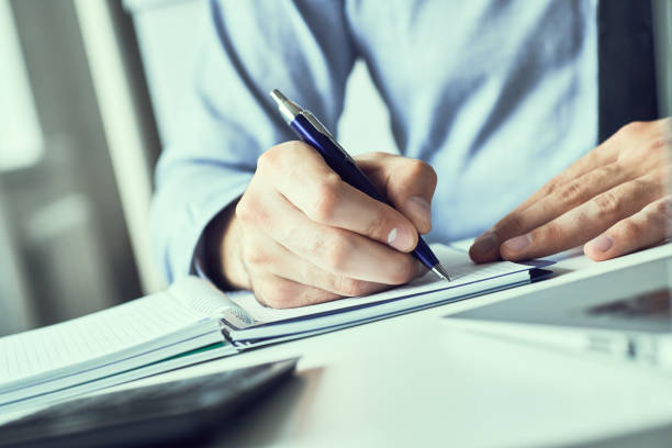 istock Businessman hand writing note on a notebook. Business man working at office desk. Close up of empty notebook on a blackboard with office supplies. 1157775866