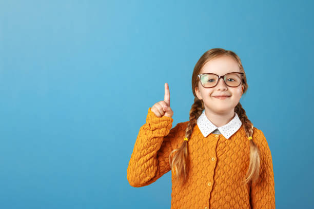 porträt einer kleinen mädchen schülerin auf blauem hintergrund. ein kind mit brille zeigt mit dem finger nach oben. kopierraum - child glasses eyewear little girls stock-fotos und bilder