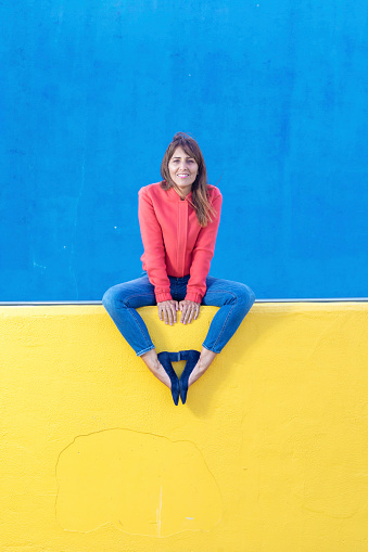 Beautiful adult woman in jeans sitting on a yellow fence while looking camera against blue wall background