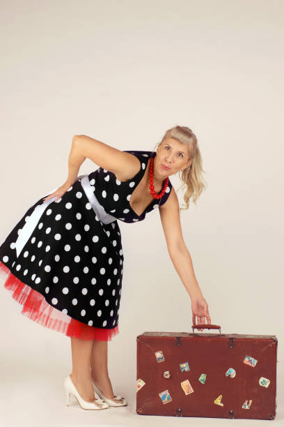 belle femme blonde dans le modèle de pin-up, rectifiée dans une robe de polka-point, se tient et soulève une valise de voyage, fond blanc - polka dot suitcase retro revival women photos et images de collection