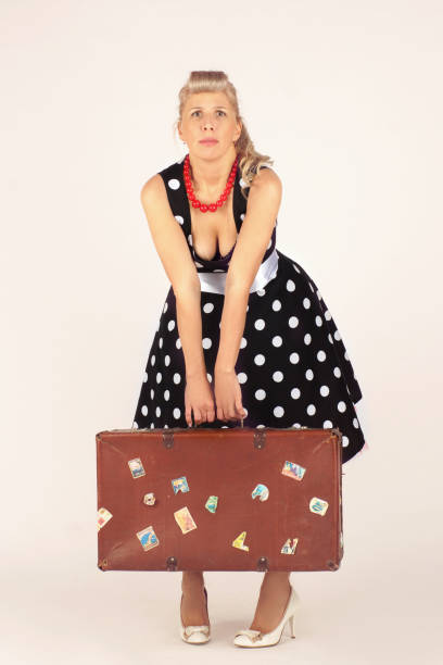 belle femme blonde dans le modèle de pin-up, rectifiée dans une robe de polka-point, se tient et tient à peine une valise lourde, fond blanc - polka dot suitcase retro revival women photos et images de collection