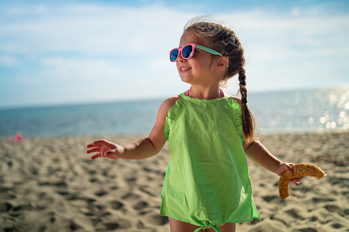 Cute little girl is standing on the beach, holding pastry and smiling.