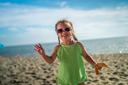 Cute little girl is standing on the beach, holding pastry and smiling.