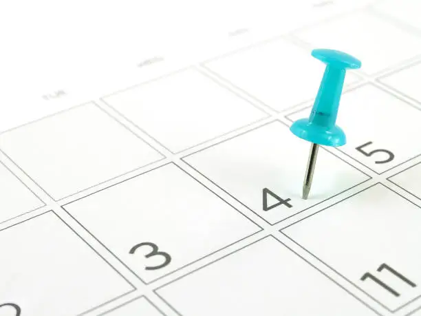 Photo of close up single blue plastic push pin tacking in schedule box of 4th of July on white desk calendar page