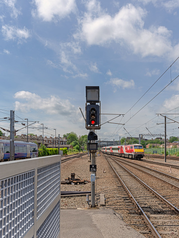 Signal light for trains at the end of a platform set to red. Two trains travel on on either side and a fence is in the foreground. A blue sky and clouds are overhead.