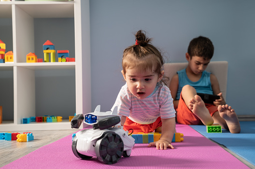 Baby girl trying to reach robot controlled by her brother. 6 years old elder brother using a mobile phone to control the robot. Baby girl is looking curiously. They are on the floor. Other toy blocks are seen around. Shot indoor with a full frame mirrorless digital camera.