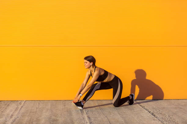 vue de côté de jeune belle femelle sportive de yogi dans le sportwear noir s'étendant sur le plancher de rue restant sur le genou, regardant droit - beautiful caucasian teenager running photos et images de collection