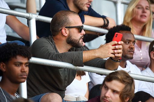 Man holding his red smart phone sitting on stadium among crowd.
