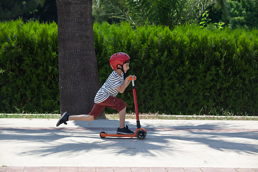 Portrait of little boy riding kick scooter on sidewalk in outdoor. He is wearing a red and white striped t-shirt and a blue short. The scooter and helmet are red. The background is a concrete high wall. Shot in outdoor daylight with a full frame mirrorless camera.