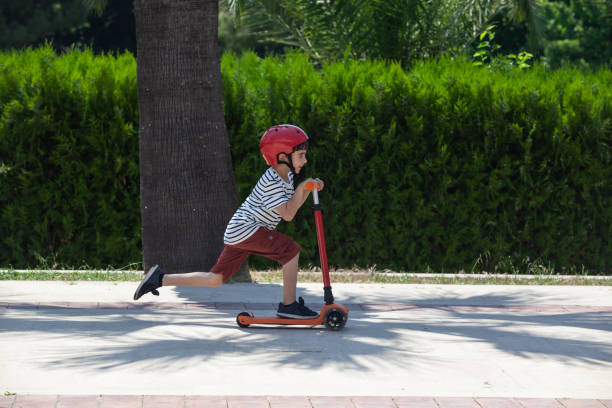 Portrait Of School Boy Riding Kick Scooter Portrait of little boy riding kick scooter on sidewalk in outdoor. He is wearing a red and white striped t-shirt and a blue short. The scooter and helmet are red. The background is a concrete high wall. Shot in outdoor daylight with a full frame mirrorless camera. push scooter stock pictures, royalty-free photos & images