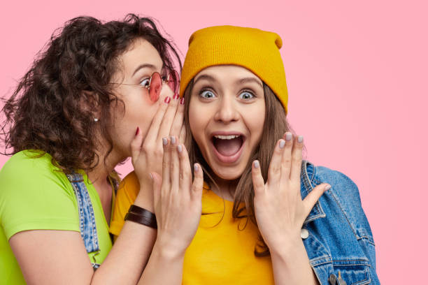 Female sharing secret with best friend Young girl in bright outfit telling secret to amazed best friend against pink background gossip photos stock pictures, royalty-free photos & images