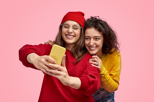 Excited female friends in bright trendy outfits smiling and taking selfie together against pink background