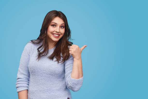 Cheerful woman pointing aside Happy female in elegant sweater friendly smiling and pointing aside with thumb while standing against blue background voluptious stock pictures, royalty-free photos & images