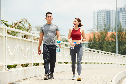 Fit smiling Asian man and woman walking on bridge with water bottles after morning run