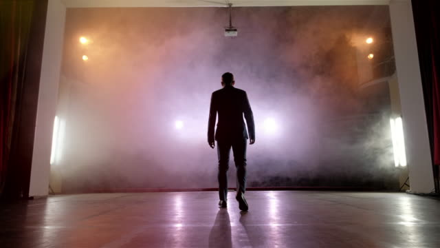 Showman. Young male entertainer, presenter or actor on stage. Back, arms to sides, smoke on background of spotlight.