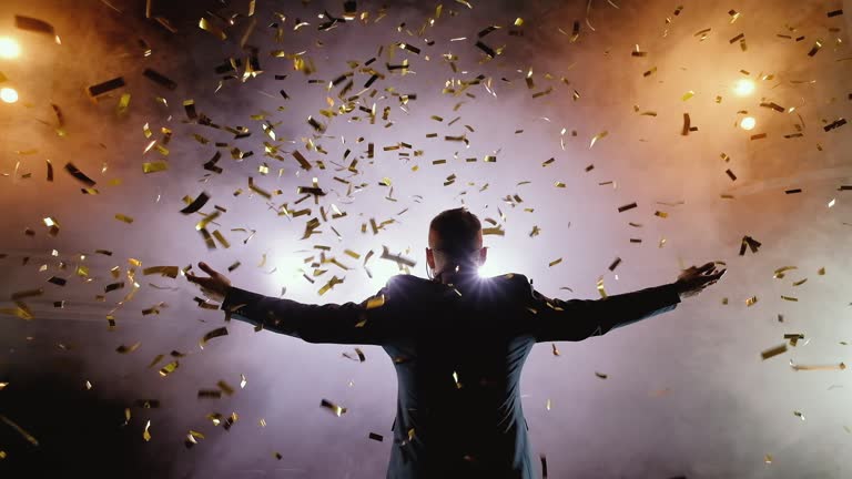 Successful businessman with arms up celebrating his victory. Celebrating success. Low angle view of excited young businessman keeping arms raised and expressing positivity while stands on the stage in the background of the hall