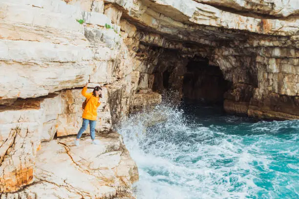 Photo of woman in yellow raincoat standing on the cliff looking into grotto