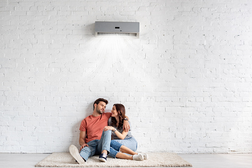 happy young couple hugging while sitting on floor near white wall under air conditioner