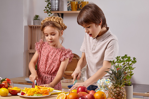 Cute kids are cooking together in a kitchen against a white wall with shelves on it.