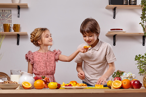 Cute kids are cooking together in a kitchen against a white wall with shelves on it.