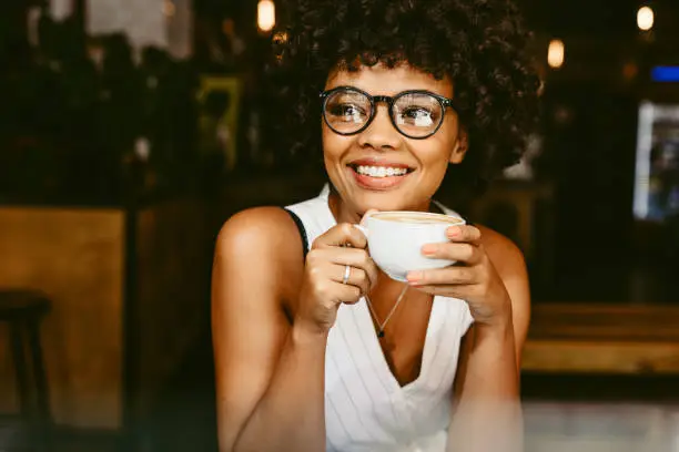 Beautiful young woman sitting at cafe with a cup of coffee and looking away smiling. African female having coffee at coffeeshop.