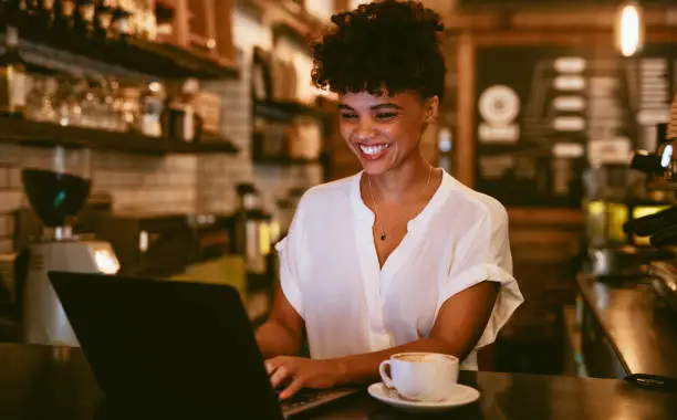 African woman standing at cafe counter working on laptop with a cup of coffee. Smiling female cafe owner using laptop.