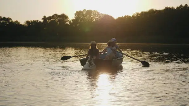 Young family floating in a boat with oars at sunset. Slow motion. Girl in a hat smiling, rowing paddles, teen daughter sitting at the stern of wooden boats and wets feet in water.