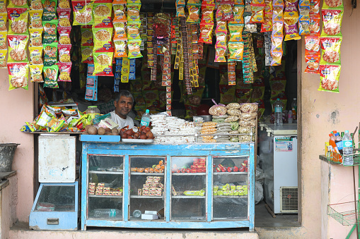 KAMBESWARJI, INDIA - 13 AUGUST 2017: Indian vendor sits in shop with chips & namkeen hanging in background and other products. pakazing namkeen and chips are big sale in india